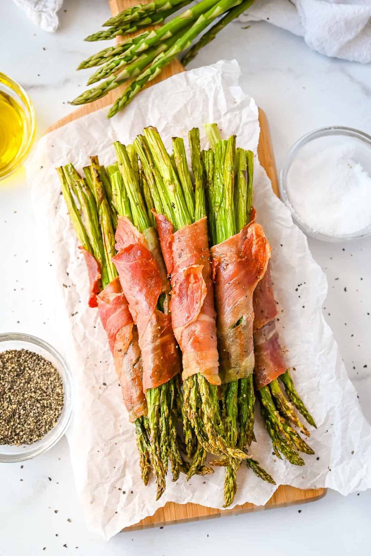 far shot of bacon wrapped asparagus on white parchment paper with salt, pepper, olive oil and asparagus.