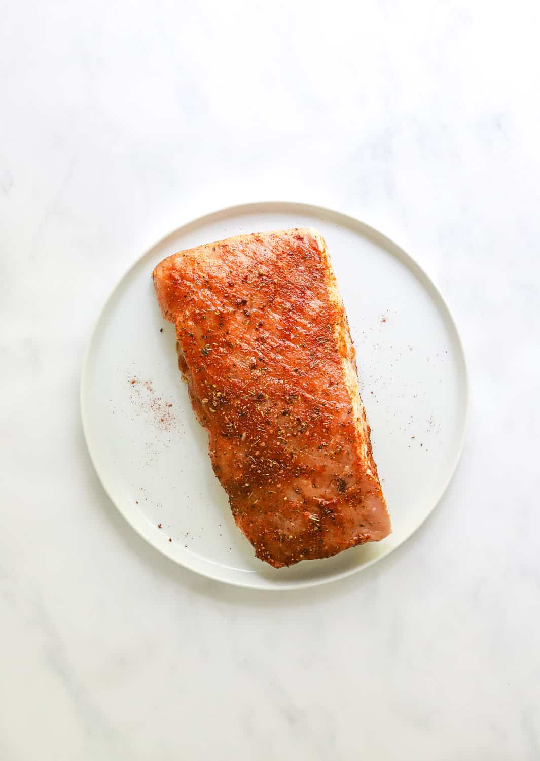 Uncooked pork loin covered in seasoning on a round, white plate.