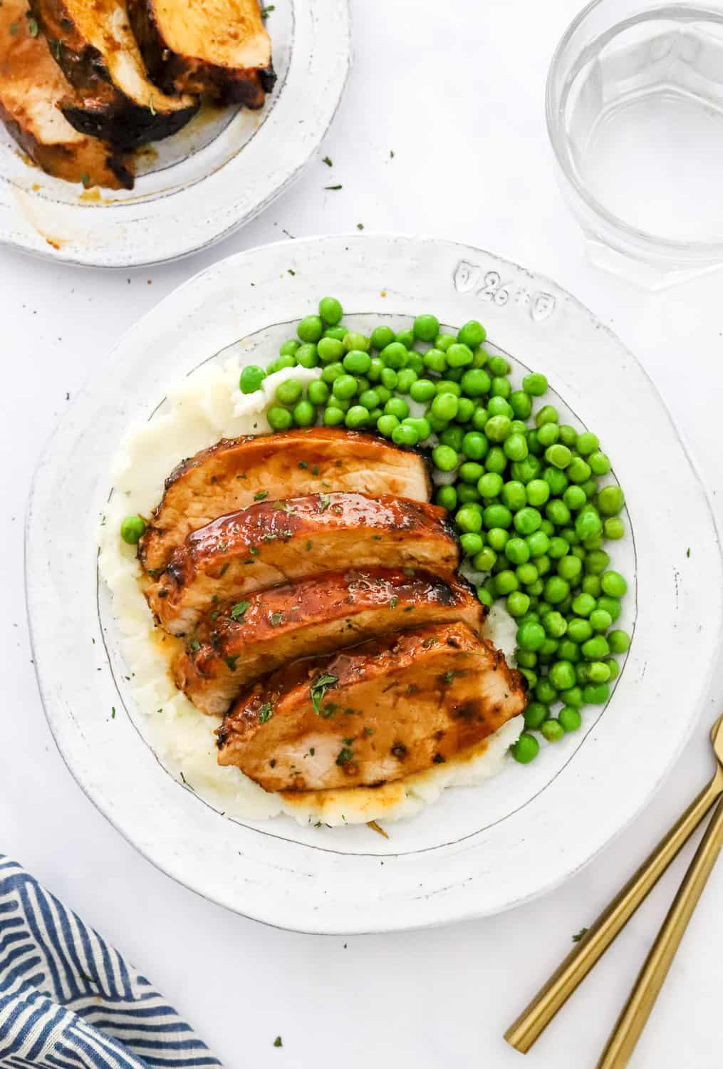 Round plate with sliced brown meat on it on top of mashed potatoes with peas on the plate with another plate of meat and glass of water behind it.