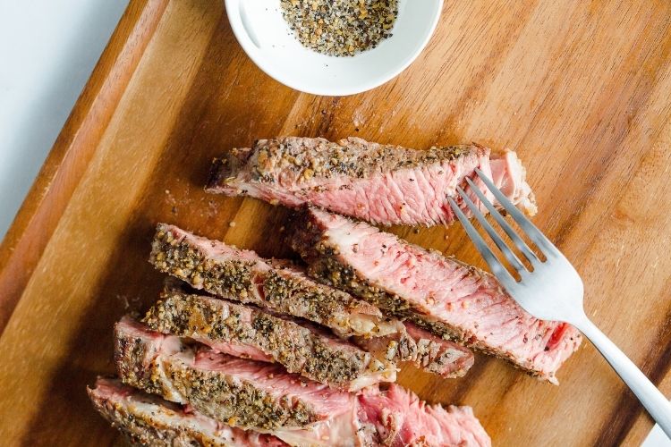 Sliced air fryer steak on a cutting board with fork and bowl of seasoning